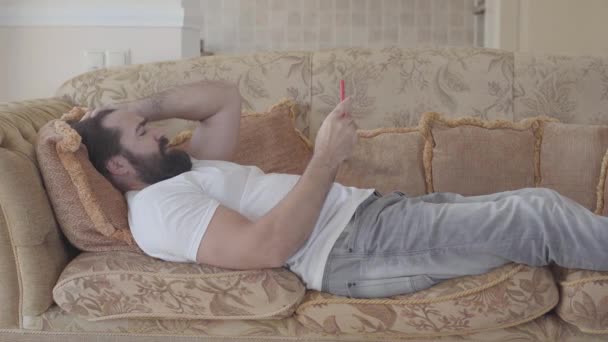 Bearded man lying on the sofa checking cell phone. Beardie is angry, he throws away smartphone. Receiving bad news. Emotional man rests at home — Stock Video