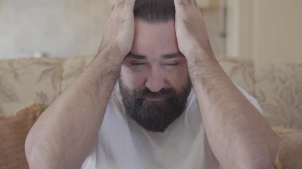 Портрет бородатого, массирующего голову большими руками с выражением страдания на лице. Взрослый мужчина испытывает сильную головную боль сидя дома на диване — стоковое видео