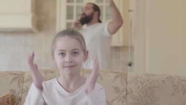 Malá dívka sedí na gauči a pokrývající uši, protože její otec je telefonujete hlasitě na pozadí — Stock video