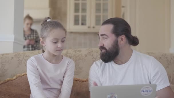 Vader met schattige dochter en laptop in de woonkamer op de bank zitten en praten over iets grappig op de achtergrond van de moeder — Stockvideo