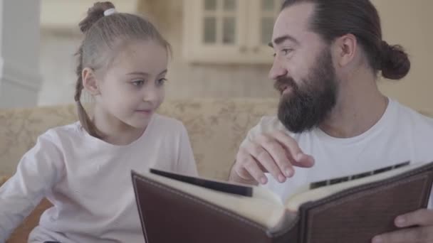 Bärtiger Mann, der auf dem Sofa sitzt und seiner kleinen Tochter das Buch aus nächster Nähe zeigt. Konzept der glücklichen Familie. kleines Mädchen verbringt Zeit mit ihrem Vater — Stockvideo