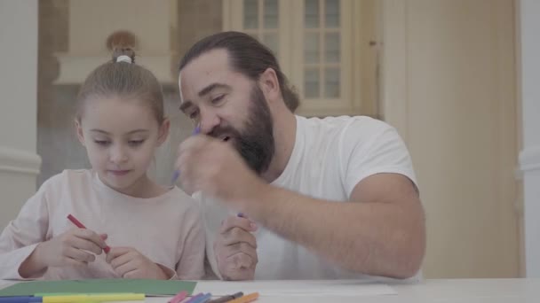 Hombre barbudo y niña dibujan con marcadores sentados a la mesa en casa. Padre ayudando a su hija a dibujar, apoyar su talento. La gente respira en el marcador para que dibuje mejor — Vídeos de Stock