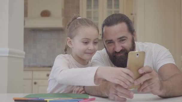 El joven padre y su pequeña hija linda están sentados en la sala de estar y buscando en la imagen del teléfono inteligente para dibujar con marcadores — Vídeos de Stock