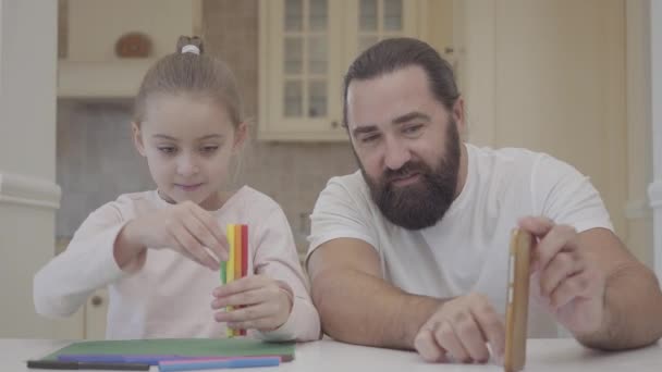 Jonge vader en zijn kleine schattige dochter zitten in de woonkamer en letten op een experiment met markeringen en gooi ze op tafel — Stockvideo
