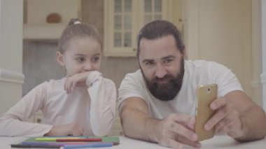 Sakallı adam küçük kız bilgi onun cep telefonu, oturmak için gösterilen mutfak tabloyu kapatın. Baba ve kızı birlikte gülüyor. Mutlu aile kavramı. Küçük kız baba ile eğlenin