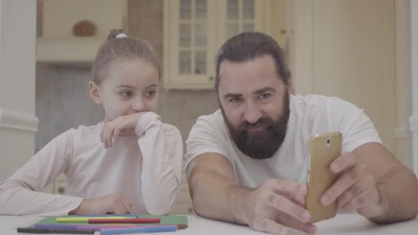 Бородатий чоловік показує маленькій дівчинці інформацію на мобільному телефоні, сидячи за столом на кухні крупним планом. Батько і дочка сміються разом. Концепція щасливої сім'ї. Маленька дівчинка розважається з татом — стокове відео