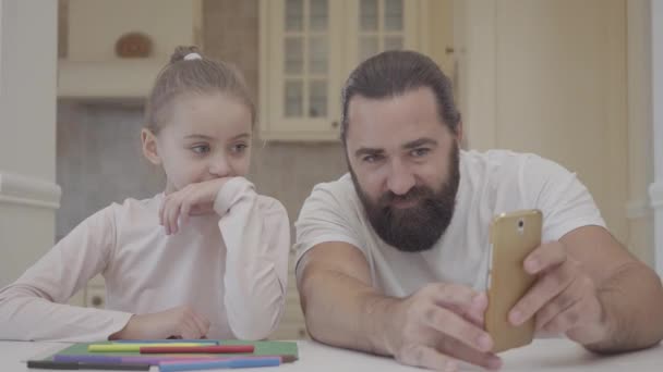 Barbudo hombre mostrando a la niña pequeña información en su teléfono celular sentado en la mesa en la cocina de cerca. Padre e hija riéndose juntos. Concepto de familia feliz. Pequeña chica divertirse con papá — Vídeo de stock