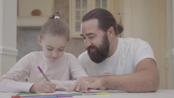 El joven padre y su pequeña hija linda están sentados en la sala de estar y dibujando con marcadores en cartón verde — Vídeos de Stock