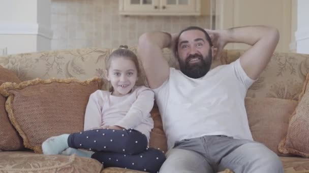 Linda hija pequeña con su padre divertido viendo una película interesante en la televisión con grandes emociones en la acogedora sala de estar — Vídeos de Stock