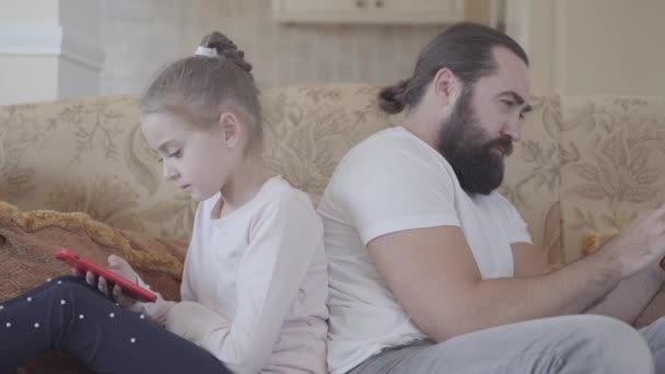 Fille mignonne et son père assis sur le canapé sur le salon avec des téléphones intelligents et se refroidissant dans une journée confortable. Relation père-fille . — Video