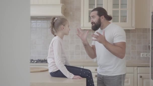 Homem barbudo contando emocionalmente história para sua filhinha, gesticulando ativamente na cozinha. Menina se senta na mesa ouvindo atentamente, em seguida, coloca o dedo nos lábios, pedindo silêncio — Vídeo de Stock