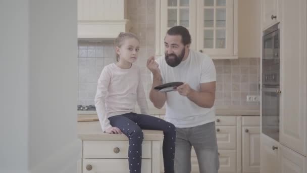 Pai emocional repreendendo sua filha por algo segurando um prato em suas mãos, quando ela está sentada chateada na mesa. Relacionamento pai e filha . — Vídeo de Stock