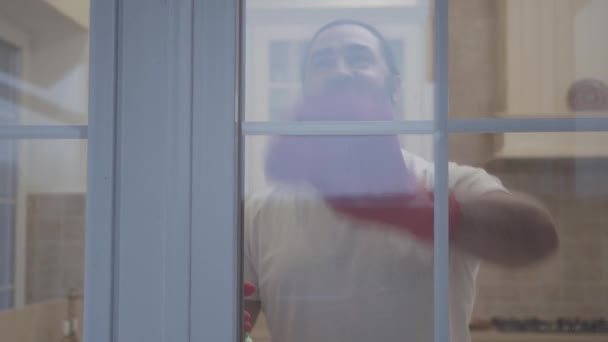 Красивий чоловік з гіпсовою красивою бородою прибирає вікно в рукавичках з миючим засобом у своєму новому сучасному будинку — стокове відео