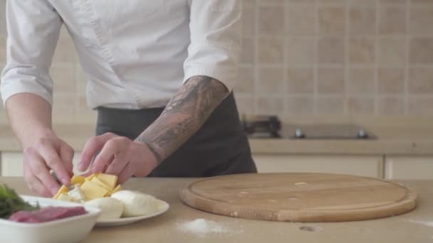 Primo piano mani di giovane pizzaiolo in grembiule che prepara prodotti per la pizza a casa. Diversi ingredienti sdraiati sul tavolo vicino al piatto di legno. Concetto di preparazione del cibo. Telecamera spostamenti a sinistra destra — Video Stock