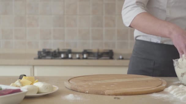 Primo piano mani di giovane pizzaiolo in grembiule preparare la pasta per la pizza a casa. Diversi ingredienti sdraiati sul tavolo vicino al piatto di legno. Concetto di preparazione del cibo. Macchina fotografica si muove a destra — Video Stock