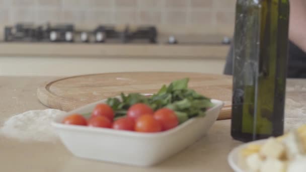 Le mani vicine di pizzaiolo giovane in uniforme di cuoco abilmente inginocchiandosi la pasta per pizza in cucina moderna. Olio d'oliva, pomodori e formaggi in tavola. Concetto di preparazione del cibo. Camera si muove a sinistra — Video Stock