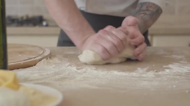 Le mani vicine di pizzaiolo giovane in uniforme di cuoco abilmente inginocchiandosi la pasta per pizza in cucina moderna. Olio d'oliva, pomodori e formaggi in tavola. Concetto di preparazione del cibo. Camera si muove a sinistra — Video Stock