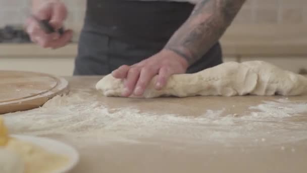 Närbild av händerna på unga pizzabagare i kock enhetlig skära deg för pizza med stor kniv i moderna kök. Olivolja, tomater basilika och ost på bordet. Kameran flyttar vänster — Stockvideo