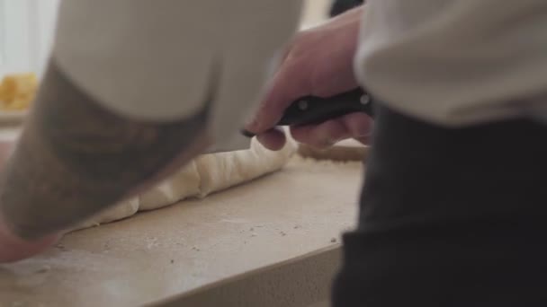 Закройте руки молодому изготовителю пиццы в форме повара, нарезая тесто для пиццы большим ножом на современной кухне. Оливковое масло, томаты базилик и сыр на столе. Камера движется влево — стоковое видео