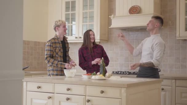 Der junge Pizzabäcker jongliert in der Küche mit Teigbällen und demonstriert seinen Freunden sein Können. lächelnder Mann und Frau, die erfolglos versuchen, seine Handlungen zu wiederholen. Pizzakunst — Stockvideo