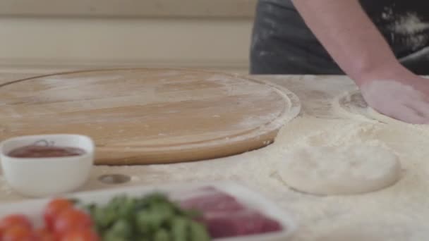 关闭年轻的比萨饼制造商在厨师制服的手形成一个比萨饼外壳揉面团在厨房。披萨食材躺在前台的桌子上。食品准备。相机向右移动 — 图库视频影像
