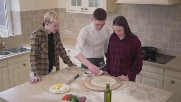 Junger Pizzabäcker bringt seinen Freunden bei, wie man zu Hause in der Küche Pizza zubereitet. Selbstbewusster Pizzaiolo, der Sahne und Tomatensauce auf den Teig gibt. Pizzakunst. Draufsicht, Schießen von oben — Stockvideo