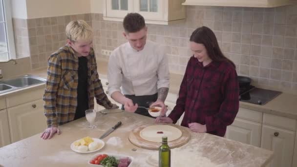 Joven fabricante de pizza enseñando a sus amigos cómo hacer pizza en la cocina en casa. Pizzaiolo seguro de aplicar salsa de tomate en la masa. Arte culinario de pizza. Vista superior, disparando desde arriba — Vídeos de Stock