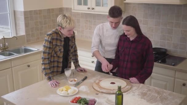 Joven fabricante de pizza enseñando a sus amigos cómo hacer pizza en la cocina en casa. El pizzaiolo seguro ayuda a la muchacha a extender la salsa de tomate en la masa. Arte culinario de pizza. Vista superior, disparando desde arriba — Vídeos de Stock