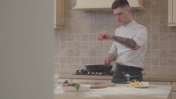 Un joven con uniforme de chef pone sal en la sartén y vomita carne en una sartén. Cocinero profesional cocinando comida en casa en la cocina — Vídeos de Stock