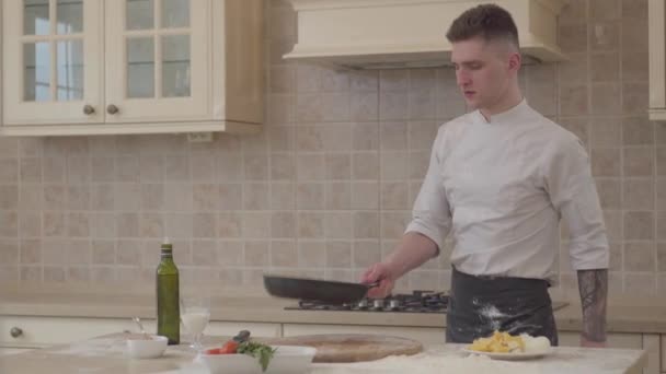 Ein junger Mann in Kochuniform legt Salz in die Pfanne und wirft das Fleisch in einer Pfanne in die Pfanne. ein professioneller Koch gießt Milch in Zutaten für Pizza und stellt die Pfanne auf den Herd. — Stockvideo