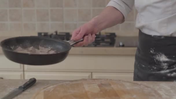 Un hombre irreconocible con uniforme de chef pone sal en la sartén y vomita carne en una sartén. Cocinero profesional cocinando comida en casa en la cocina — Vídeo de stock