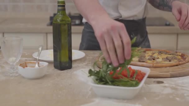 Un primer plano de manos de hombre irreconocibles pone rúcula en la parte superior de su pizza acostada sobre la mesa. Concepto de preparación de alimentos. Cámara se mueve derecha — Vídeo de stock