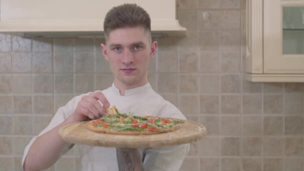 Portrait of attractive smiling young man holding wooden board with pizza close up. Concept of food preparation. Professional cook cooking pizza at home in the kitchen. Camera moves left — Stock Video