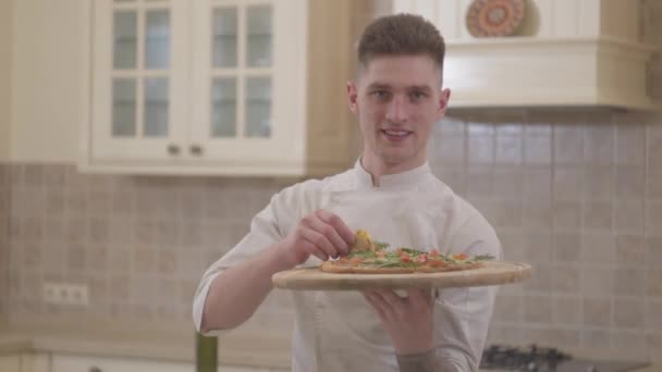 Portret van knappe lachende jonge man houden van houten plaat met pizza close-up. Concept van voedselbereiding. Professionele kok koken pizza thuis in de keuken. — Stockvideo