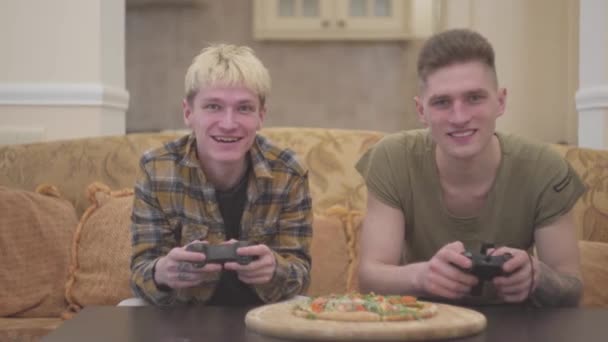 Δύο φίλοι κάθονται στον καναπέ στο σαλόνι και παίζει video game με ενθουσιασμό χαμογελώντας εκμετάλλευση πηδάλιο στα χέρια. Πίτσα που βρίσκεται κοντά στο τραπέζι. Ελεύθερου χρόνου των φοιτητών τους φίλους — Αρχείο Βίντεο