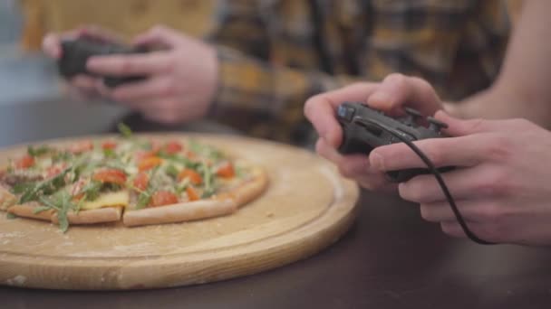 Κλείστε τα χέρια των δύο νεαροί άνδρες που παίζει video game χειριστηρίου στα χέρια στο σπίτι. Πίτσα με ντομάτες και ρόκα που βρίσκονται σε πρώτο πλάνο στο τραπέζι. Αναψυχής και των φίλων — Αρχείο Βίντεο