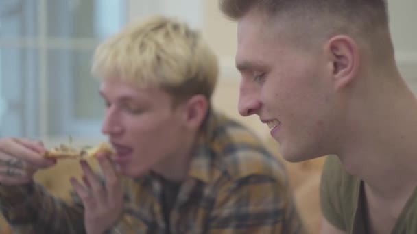 おいしい肉とルッコラのピザを食べるリビング ルームのソファに座っている二人の若者をクローズ アップ。友人の学生のレジャー。一人の男がぼやけています。 — ストック動画