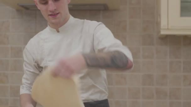 Jonge kerel in uniform van de chef-kok spinnen en woelen schijf van pizza deeg in keuken thuis. Professionele pizzaiolo pizza maken. Concept van voedselbereiding — Stockvideo