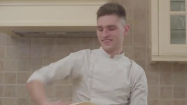 Junger lächelnder Kerl in Kochuniform, der zu Hause in der Küche eine Scheibe Pizzateig dreht. professionelle Pizzaiolo-Herstellung von Pizza. Konzept der Lebensmittelzubereitung — Stockvideo