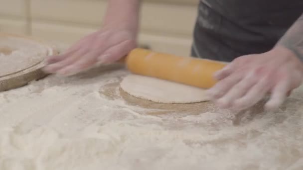 关闭年轻的比萨饼制造商的手在围裙准备面团使用面团滚针在家里在厨房。食品制备的概念。相机向右和向左移动 — 图库视频影像