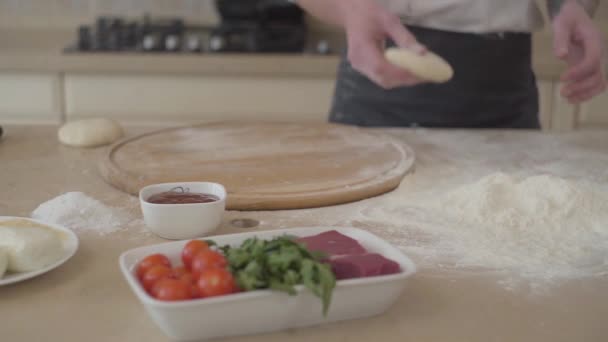 Le mani vicine di pizzaiolo giovane in uniforme di cuoco lanciano la palla di pasta nella cucina. Ingredienti della pizza sdraiati sul tavolo in primo piano. Preparazione del cibo. La telecamera si muove a destra. Rallentatore — Video Stock