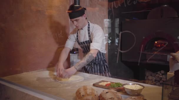 El hábil fabricante de pizza está cocinando masa en la cocina moderna del restaurante detrás del vidrio. El tipo pone los ingredientes de la pizza en la masa. Proceso de cocción de pizza — Vídeos de Stock