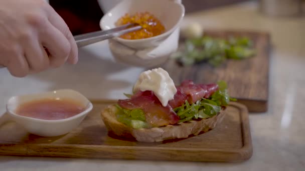 Mãos cozinheiros decorando delicioso sanduíche com caviar vermelho. Aperitivo de pão delicioso com peixe vermelho, creme e caviar vermelho em uma placa de cozinha de madeira close-up . — Vídeo de Stock