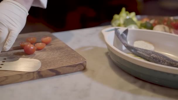 As mãos de Chef em luvas de borracha brancas cortam tomates cereja com a grande faca e põem-nos no boliche com peixe e abobrinha fecham. Preparação de alimentos em restaurante moderno — Vídeo de Stock
