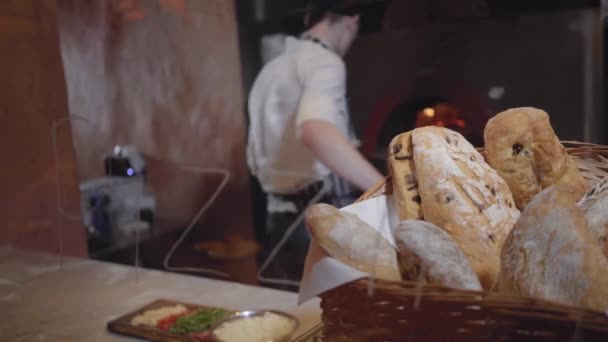 Frisch gebackenes Brot, das im Korb im Vordergrund auf dem Tisch in einem modernen Restaurant in Großaufnahme liegt. Koch und Ofen mit loderndem Feuer im Hintergrund — Stockvideo