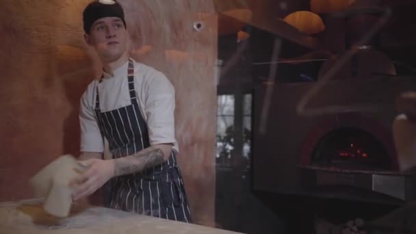 Behendig pizza maker is koken deeg op moderne restaurant keuken. Jonge kerel in chef-kok uniforme spinnen pizza deeg in restaurant keuken. Pizza koken proces. Camera omlaag — Stockvideo