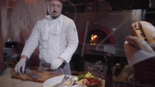 Un chef profesional con chaqueta blanca y guantes de chef de goma prepara un delicioso plato de pescado y verduras frescas. El cocinero pone los tomates en una tabla de madera y comienza a cortar . — Vídeos de Stock