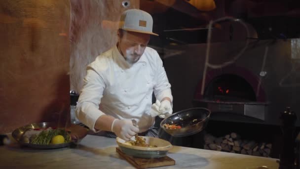 Chef em uniforme branco pondo cortado em pequenos pedaços de verduras cozidas no vapor no peixe cozido que está em chapa profunda na cozinha. Preparação de comida em restaurante moderno atrás de vidro com assinatura — Vídeo de Stock