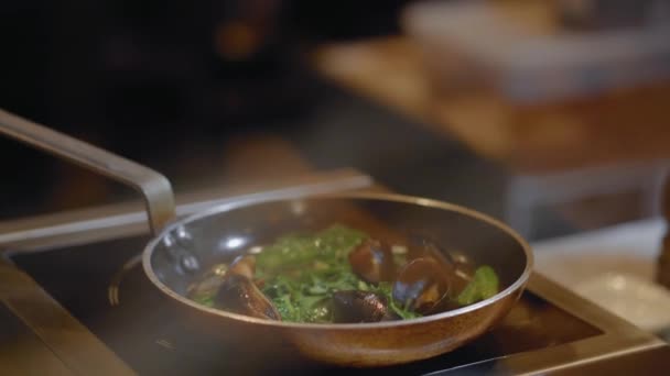 Vista de cerca de los mejillones fritos con hojas en la sartén con aceite y ajo. Preparación de mariscos en la cocina del restaurante. Fondo borroso. Cámara se mueve hacia abajo — Vídeo de stock