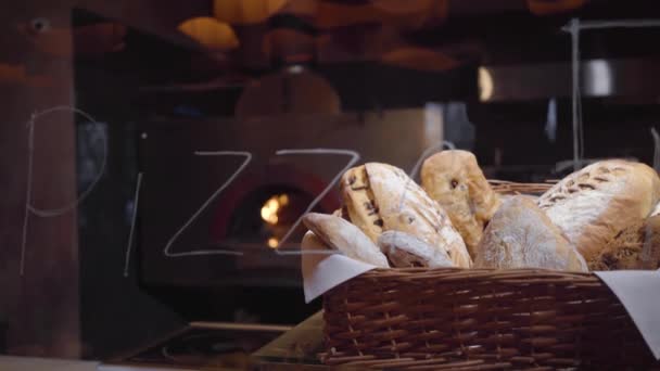 Pão recém-assado deitado na cesta em primeiro plano sobre a mesa no restaurante moderno de perto. Forno com fogo ardente no fundo. Tiro por trás do vidro com assinatura — Vídeo de Stock